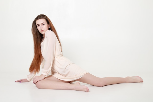 Beautiful tender young girl in a white long dress and with long hair sits in a relaxed pose in the studio on a gray background.