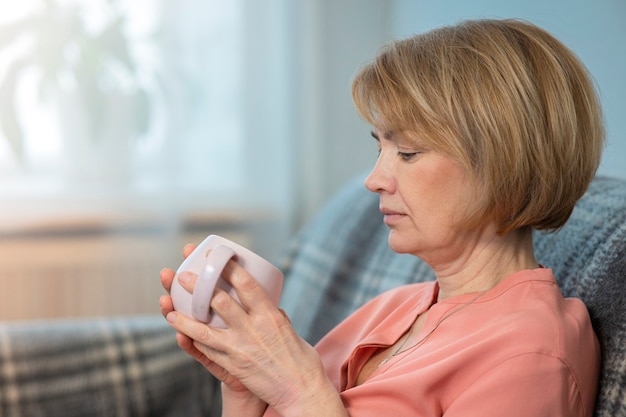 Beautiful thoughtful pensive woman is drinking tea or coffee from a cup sitting at couch at home in