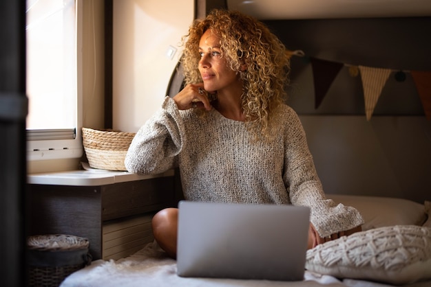 Beautiful thoughtful woman with curly hair and knitted top daydreaming or thinking of ideas, good future plans while looking out through window sitting on cozy bed with laptop at home