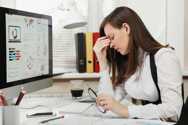 Beautiful tired perplexed and stress brown-hair business woman in suit and glasses sitting at the desk with cup of coffee, working at contemporary computer with documents in light office.