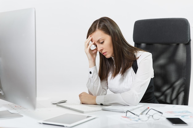 The beautiful tired perplexed and stress brown-hair business woman in suit and glasses sitting at the desk, working at contemporary computer with documents in light office