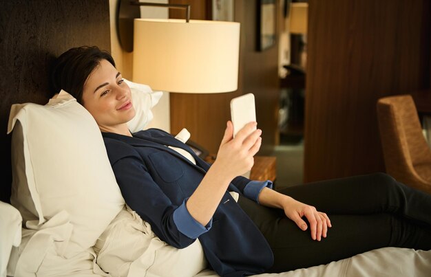 Beautiful tired woman in business suit, cutely smiles looking at camera, using a mobile phone while lying on the bed in bedroom