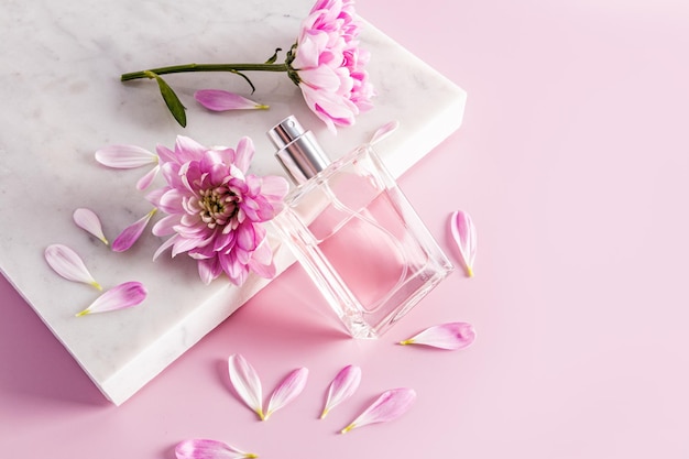 A beautiful transparent bottle with a cosmetic natural remedy spray or perfume leaned on a marble white podium with petals and flower buds