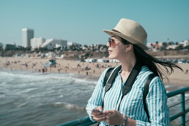 Beautiful traveler woman smiling and holding mobile phone on pier beach. young asian chinese happy lady using smartphone and enjoy beach view. female tourist holding cellphone sightseeing blue ocean