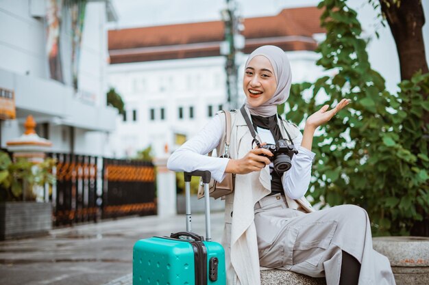 Beautiful traveller with hijab raising her opened hand while sitting on the stone seat and holding t