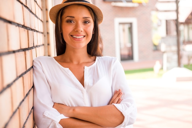 Beautiful and trendy. Beautiful young woman in hat keeping arms crossed and smiling while leaning at the wall outdoors