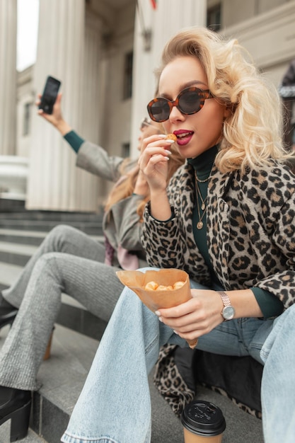 Beautiful trendy young blonde girl in fashionable vintage clothes with sunglasses sitting on the steps drinking coffee and eating cake in the city Street Style Beauty and Fashion