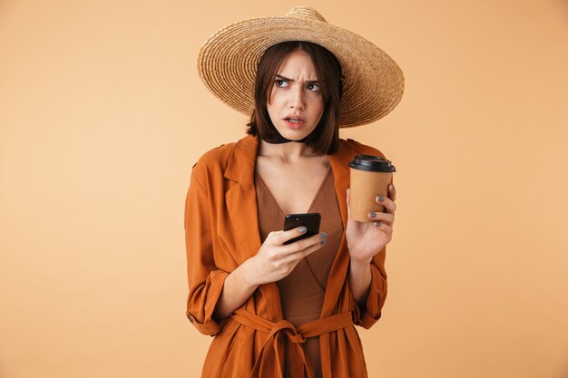 Beautiful upset young woman wearing straw hat and summer outfit standing isolated over beige wall, holding takeaway coffee cup, using mobile phone