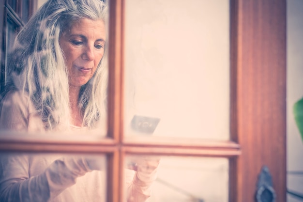 Beautiful vintage old style portrait of alternative senior woman typing and reading a mobile phone device at home viewed from the window 