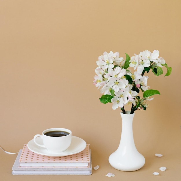 Beautiful white apple tree flowers in a white vase and notebooks for notes and a cup of tea on a beige background with copy space