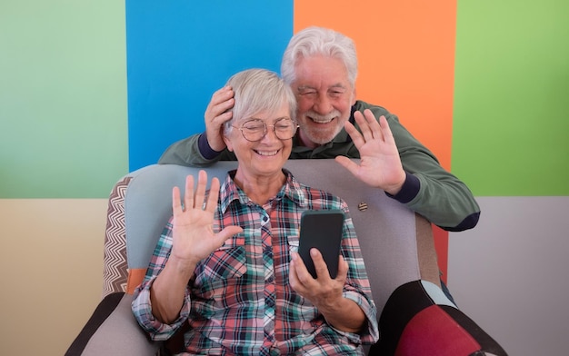 Beautiful whitehaired senior couple sitting and looking at mobile phone in video chat on colorful background Smiling elderly caucasian people relax enjoying tech and social with smart phone