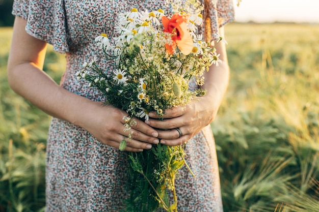 Beautiful wildflowers bouquet in woman hands close up in sunset light in barley field Stylish female relaxing in evening summer countryside and gathering flowers Atmospheric tranquil moment