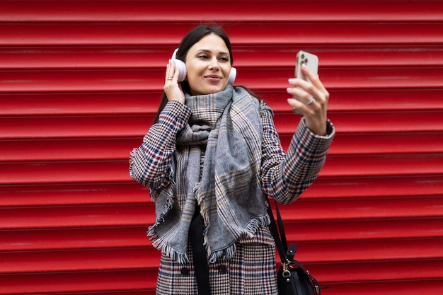 Beautiful woman in autumn clothes makes a selfie in headphones on the background of a red embossed