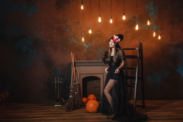 Photo a beautiful woman in a black witch costume stands near the stairs by the fireplace in the background lanterns are burning and pumpkins are lying