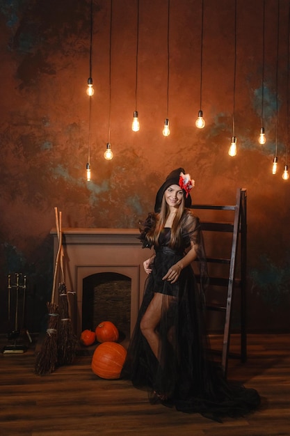 Photo a beautiful woman in a black witch costume stands near the stairs by the fireplace in the background