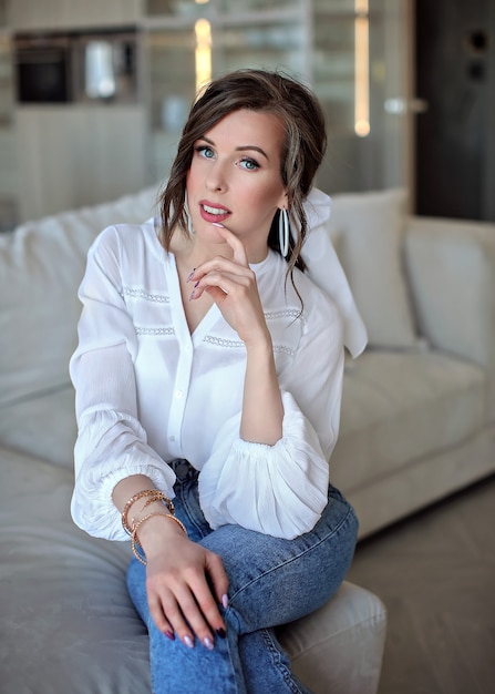 beautiful woman in blue jeans and a white shirt is sitting on the sofa. Portrait of a young woman.