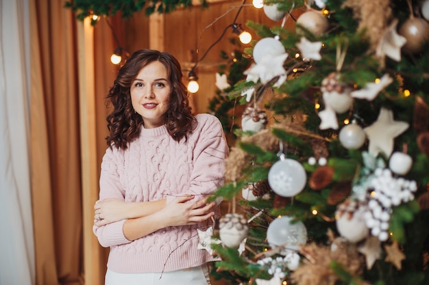 Beautiful woman brunette in pink sweater, posing near christmas tree