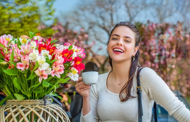 Photo beautiful woman on a cafe enjoying morning with cup of coffee she holds a cup laughing spring flowers woman drink coffee spring woman fashion concept morning coffee spring concept