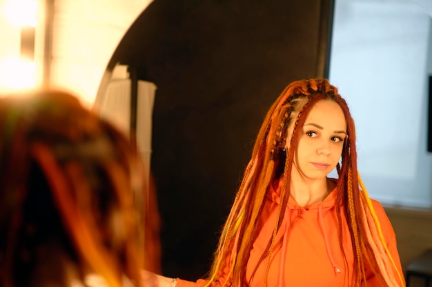 A beautiful woman in casual clothes and with dreadlocks looks in the mirror standing in a modern apartment