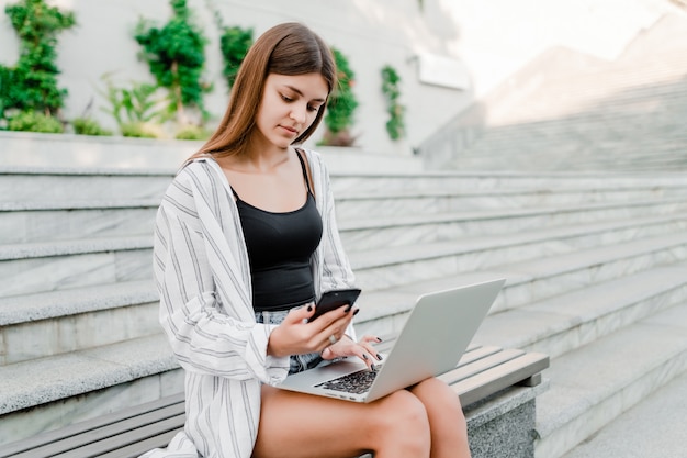 Beautiful woman checking her phone and typing on laptop outdoors