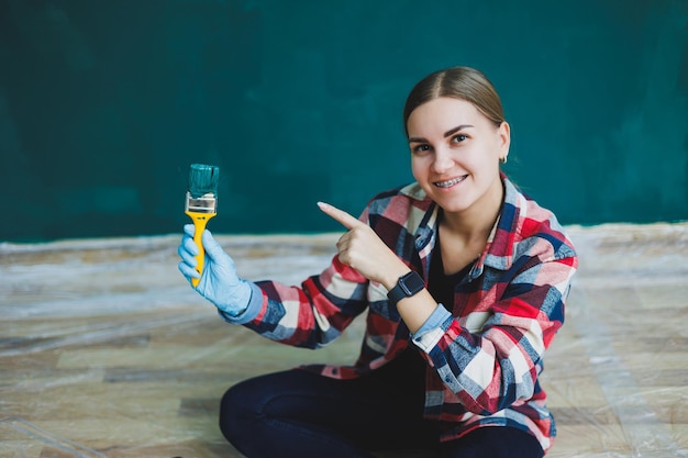 A beautiful woman designer makes repairs paints a gray wall with green paint with a brush Plaid shirt long hair and jeans Repair in the apartment