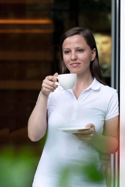 A beautiful woman drinks coffee or tea from a white mug standing in the doorway expecting friends or...