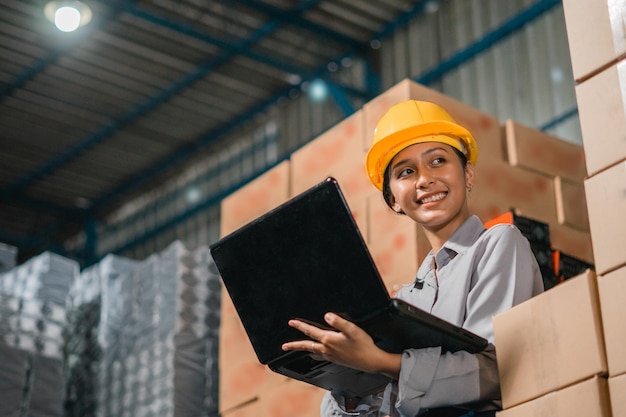 Beautiful woman factory worker wearing safety helmet while working with laptop in factory warehouse