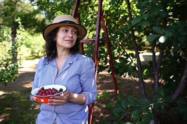 Beautiful woman farmer enjoying a summer day holding bowl with cherries freshly picked from tree in cherry orchard