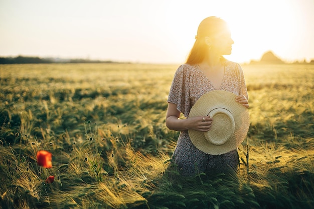 Beautiful woman in floral dress standing in barley field in sunset light Atmospheric tranquil moment rustic slow life Stylish female holding straw hat and enjoying evening summer countryside