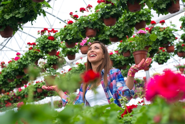 Beautiful woman florist with toothy smile holding potted flowers in plant nursery greenhouse.