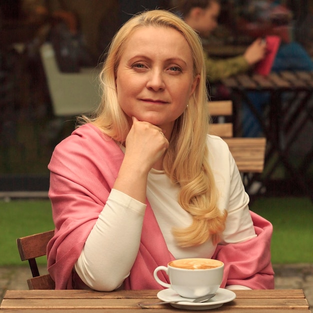 A beautiful woman forty years old dressed on a pink scar is drinking coffee in a cafe