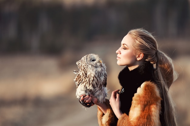 Beautiful woman in a fur coat with an owl on his arm. Blonde with long hair in nature holding a owl. Romantic delicate image of a woman