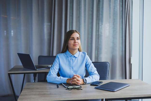 A beautiful woman in glasses sits at a table in the office and works at a laptop Corporate work in the office Modern woman workplace manager in classic clothes