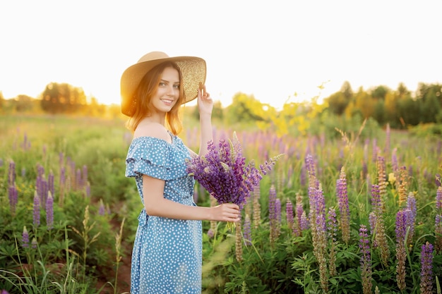 A beautiful woman in a hat and dress holds a bouquet of lupines and smiles