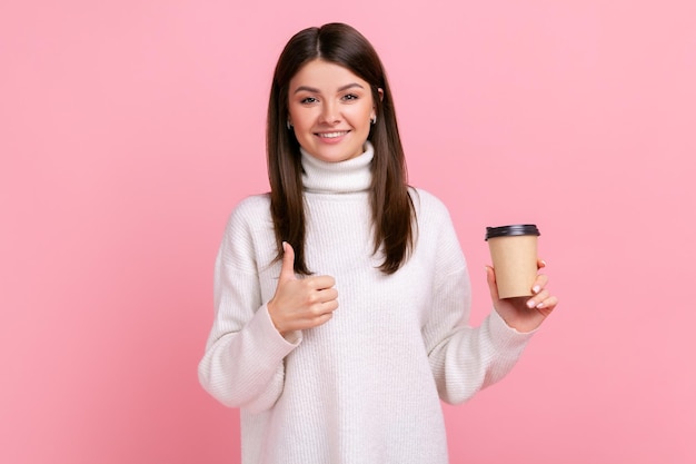 Beautiful woman holding coffee to go and showing thumb up to camera, recommend cafe or coffee house, wearing white casual style sweater. Indoor studio shot isolated on pink background.