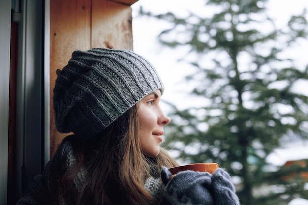 Beautiful woman holding and drinking a cup of coffee or cocoa in gloves sitting home by the window