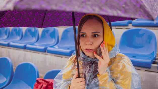 A beautiful woman in a hoodie and a plastic raincoat talking on a mobile phone sitting on the podium with an umbrella in her hand during the rain