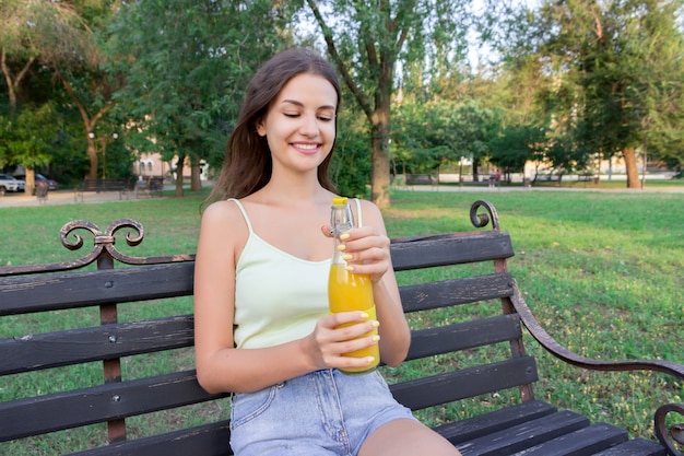 Beautiful woman is opening a bottle of fresh cold juice on the bench in the park in hot summer day