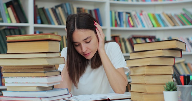 Beautiful woman is sitting at table in university library and read a book She is tired and exhausted shaking her head and writing in copybook Self education concept