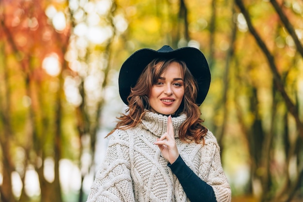 Beautiful woman in a knitted cardigan and hat touches collar by her hands in the park on the background of the autumn landscape