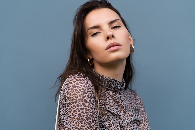 Beautiful woman in leopard fitting dress with white bag posing on blue wall background wearing golden round earrings fashion stylish