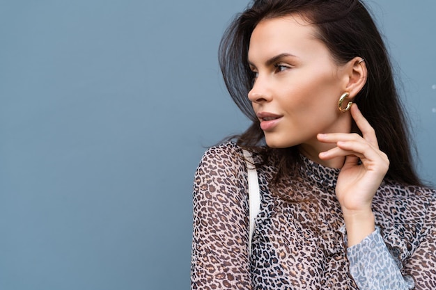 Beautiful woman in leopard fitting dress with white bag posing on blue wall background wearing golden round earrings fashion stylish