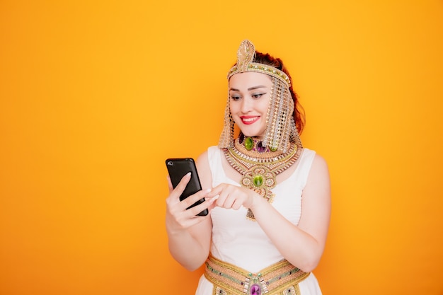 Beautiful woman like cleopatra in ancient egyptian costume holding smartphone typing a message happy and positive smiling cheerfully on orange