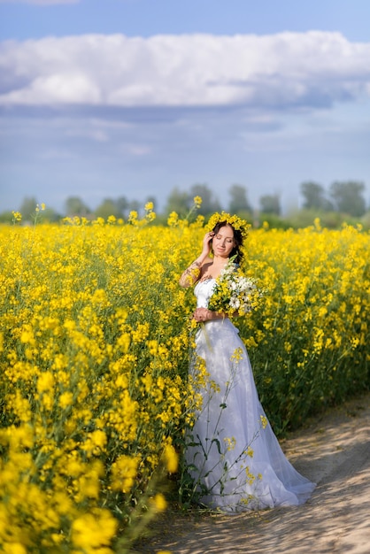 Beautiful woman in a long white dress collects flowers in a bouquet A girl against the background of a rapeseed field and a blue sky