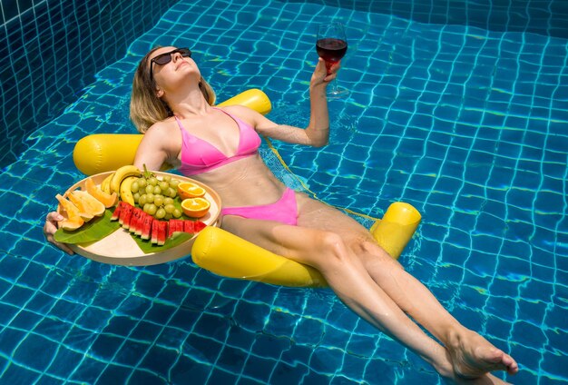 Beautiful woman lying on floating hammock in the swimming pool with wine and tray of fruits