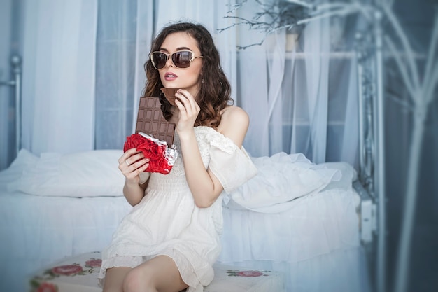 Beautiful woman model with tiles of dark chocolate in hands against bedroom interior