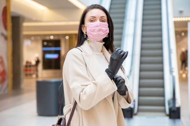 Beautiful woman in pink medical mask putting on leather gloves in a public place on background escalator