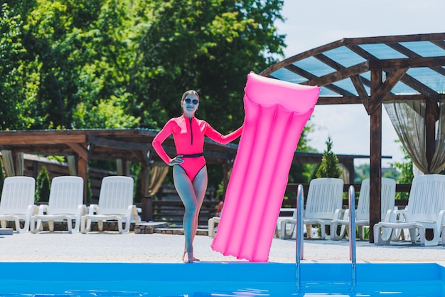 A beautiful woman in a pink swimsuit and with an inflatable mattress stands near a large pool Summer holidays in a hotel with a swimming pool Young girl in swimsuit and sunglasses