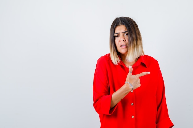 Beautiful woman pointing right in red blouse and looking puzzled. front view.