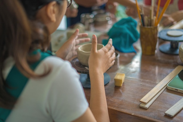 Beautiful woman potter working on potters wheel making ceramic pot from clay in pottery workshop Focus hand young woman attaching clay product part to future ceramic product Pottery workshop